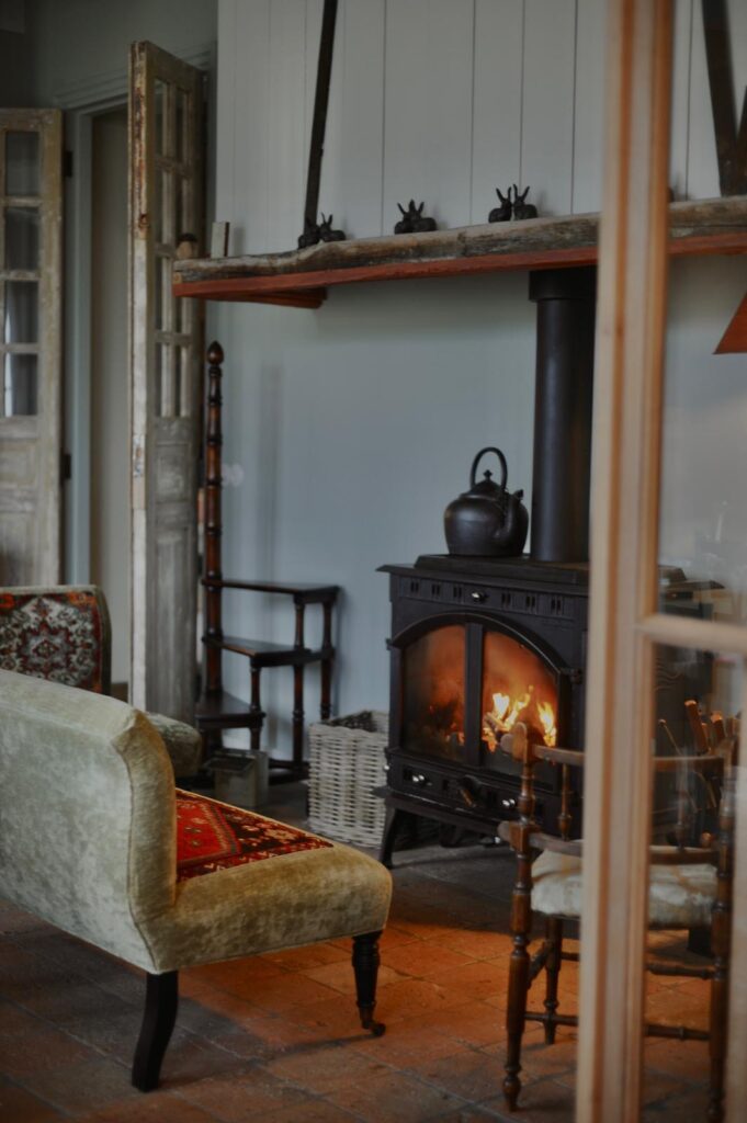 Cozy living room with a glowing wood stove, wooden beam shelf, and vintage furniture.