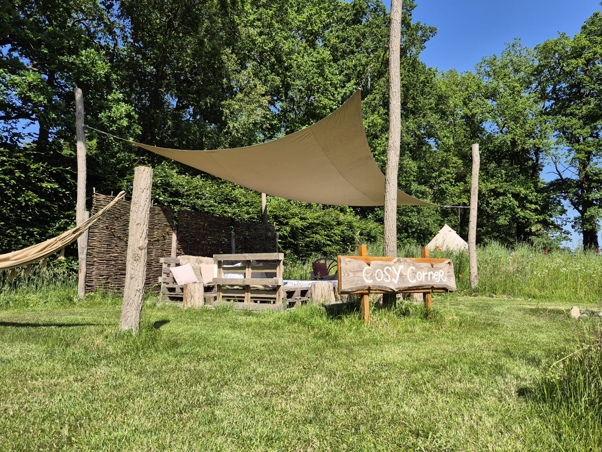 Outdoor rustic seating area under a beige shade sail between wooden posts, with a 'Cosy Corner' sign in front. Trees and blue sky in the background.