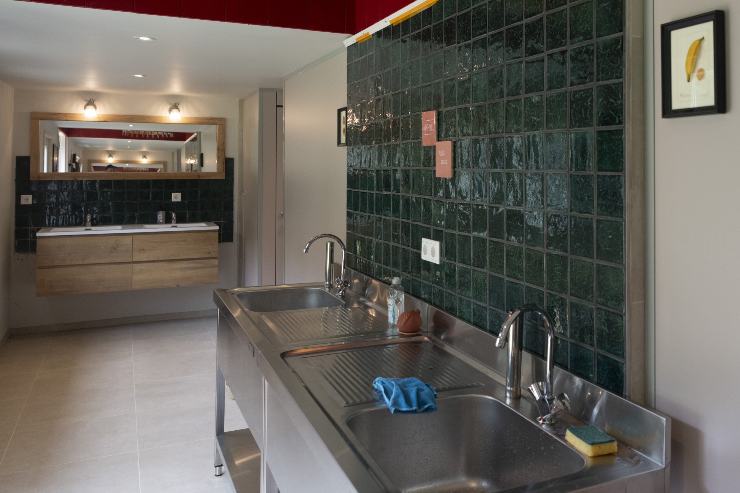 Modern kitchen with a stainless steel sink island, green tiled wall, and a large wooden-framed mirror reflecting lights.