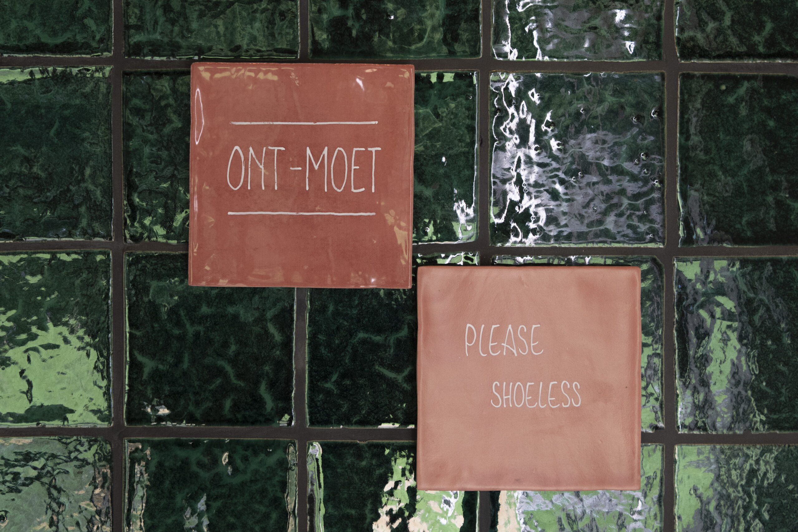 Orange ceramic tile with handwritten white text reading 'SHOELESS' on a green glass tile wall.