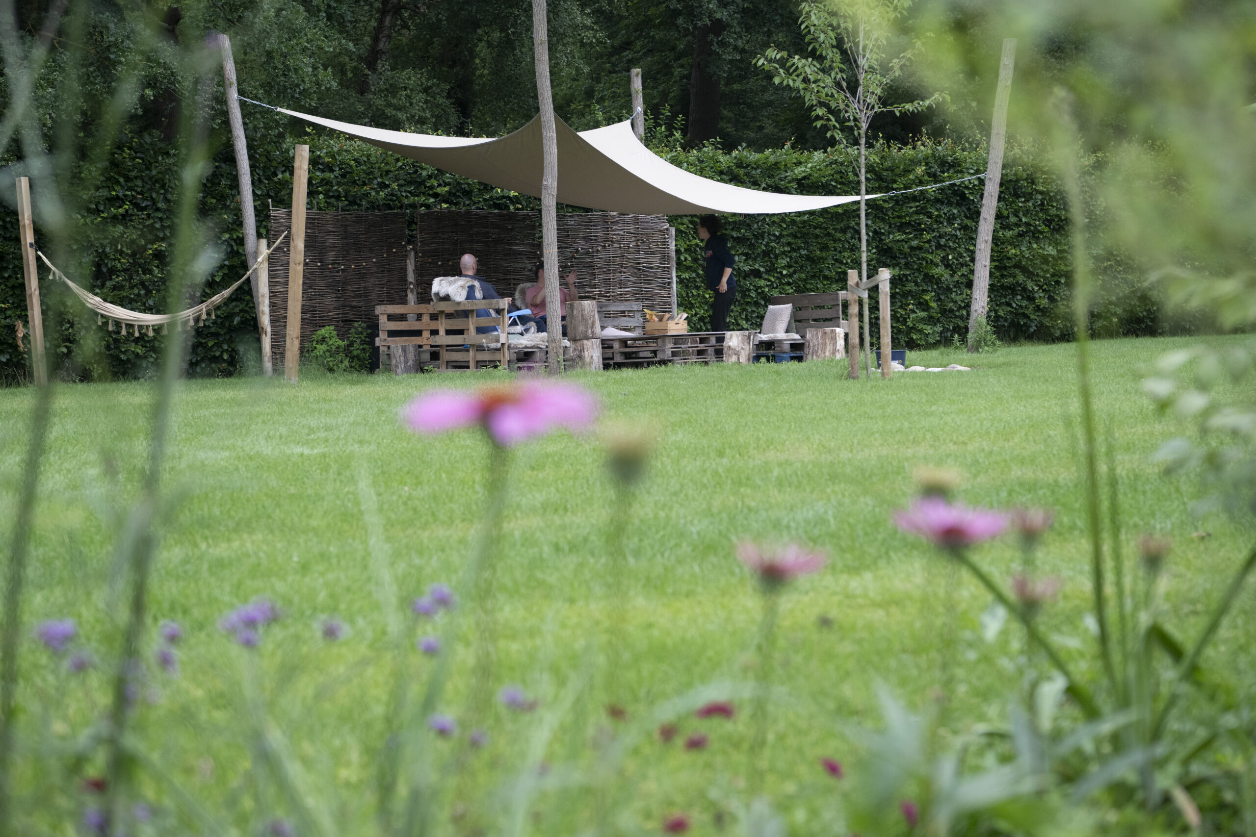 People sit and chat under a beige shade sail in a rustic garden seating area with wooden pallets and a hammock nearby.