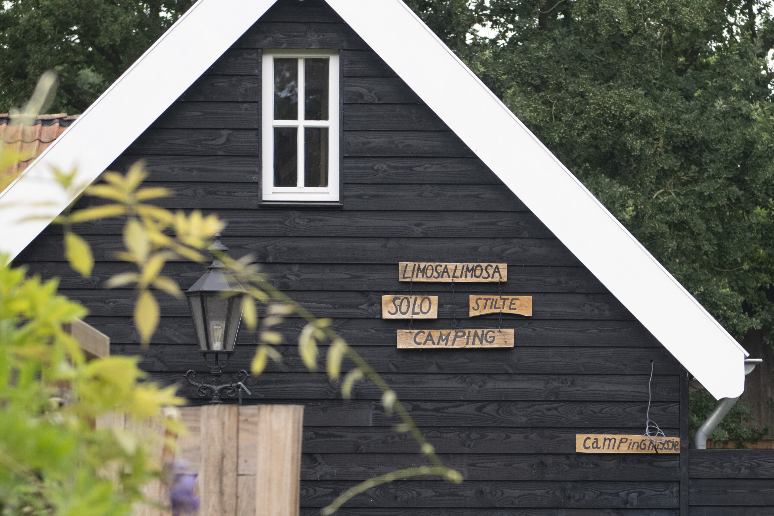 Black wooden cabin with a white-trimmed triangular roof, a small window, lantern, and wooden signs about camping on the wall.