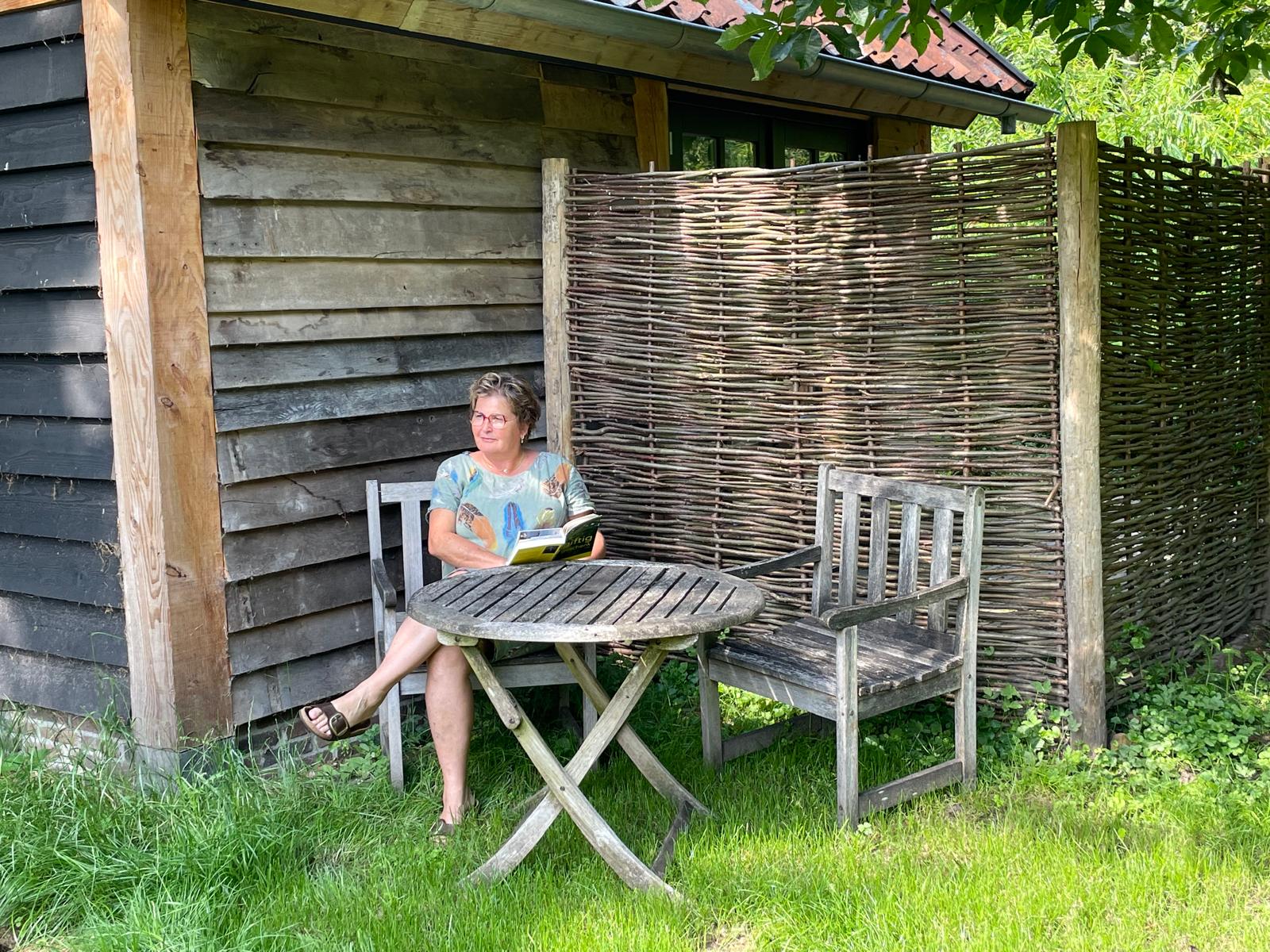 Woman sitting at a weathered wooden table outdoors, reading a magazine beside a rustic shed and wicker screen fence.