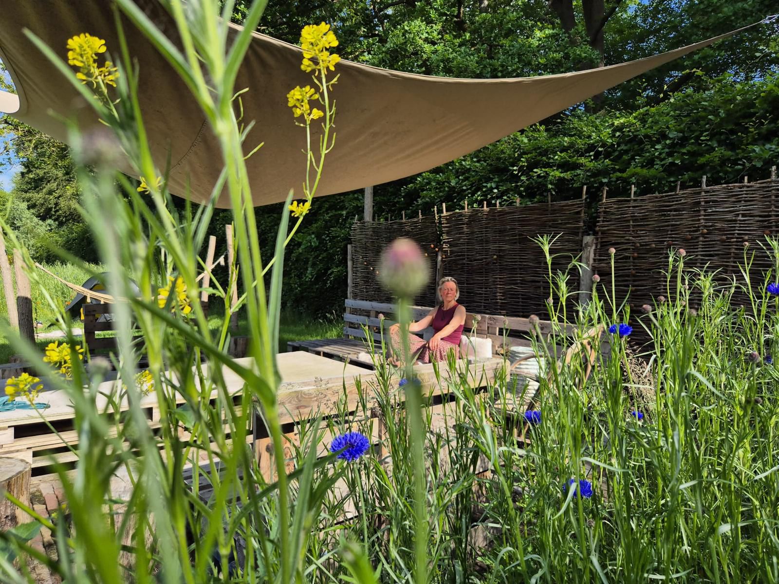 Woman sitting on a wooden deck in a lush garden under a tan shade sail, with tall yellow flowers in the foreground.