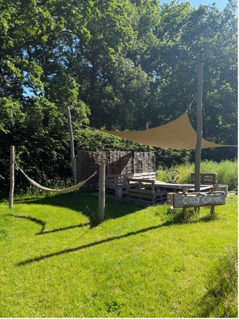 Cozy outdoor seating area under a beige shade sail, with a hammock and a 'Cosy Corner' sign in a sunny garden.