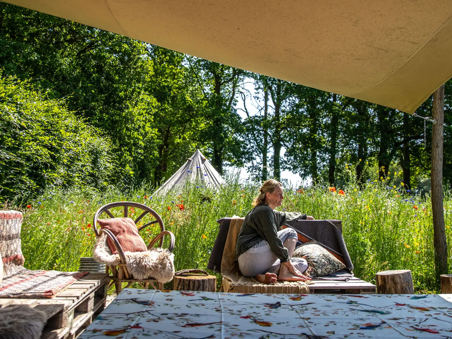 Woman seated on cushions in a sunny garden camp with a small conical tent in the background and a beige canopy above.