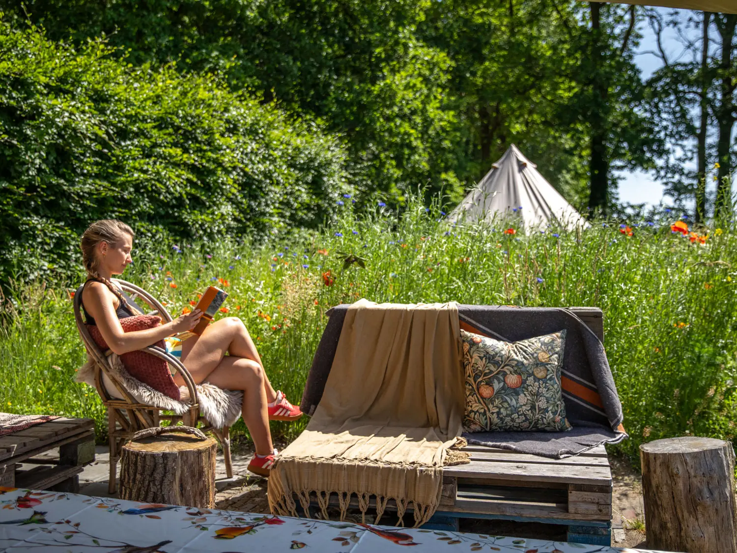 Woman relaxing in a wicker chair outdoors, reading a book, with a beige throw and floral pillow on a pallet sofa amid tall wildflowers and trees.