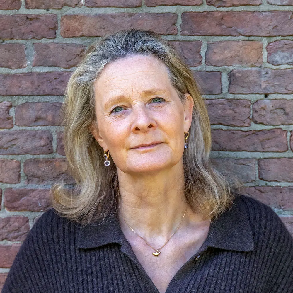 Head-and-shoulders portrait of a woman with shoulder-length gray-blonde hair, standing in front of a red brick wall, wearing a dark knit shirt and gold jewelry.