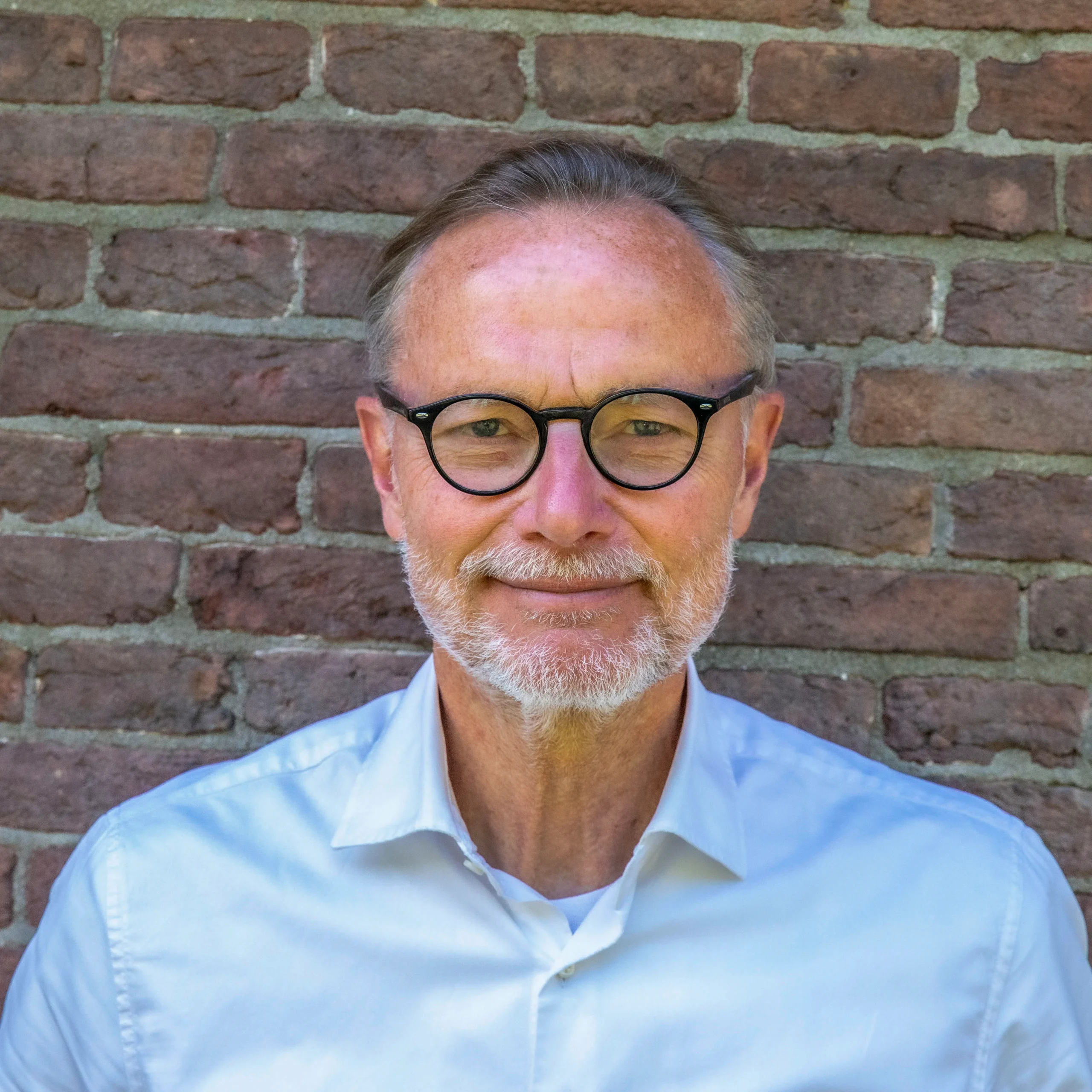 Close-up of a man with gray hair and a beard wearing black-rimmed glasses against a brick wall in the background.