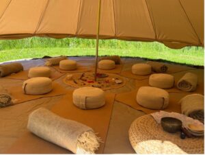 Inside a large beige tent, a circular seating area with beige cushions and rolled blankets around a central fire pit ring, on an orange mat.