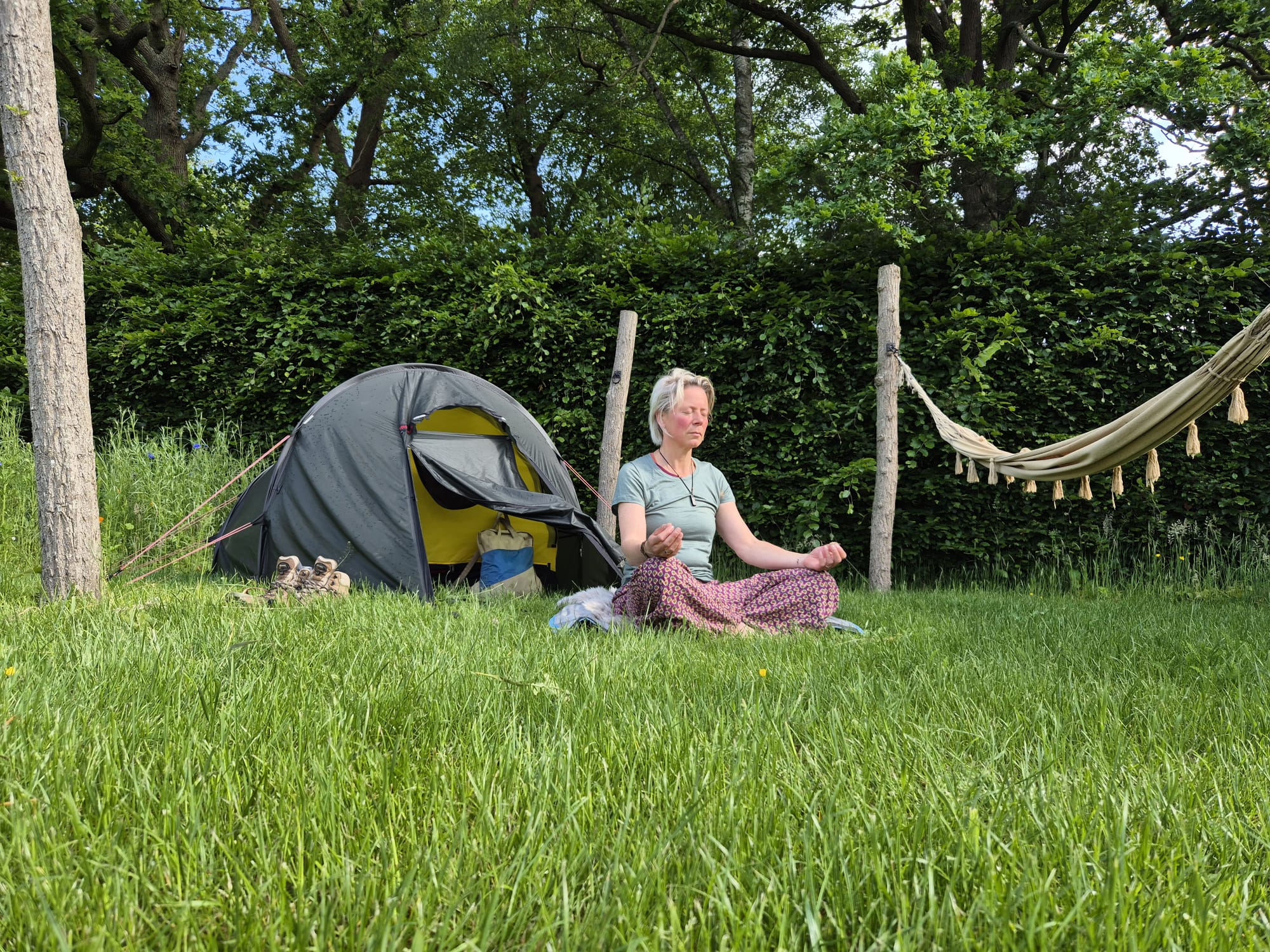 Person meditating on grass at a campsite with a gray tent nearby and a beige hammock between wooden posts in a green, leafy setting.
