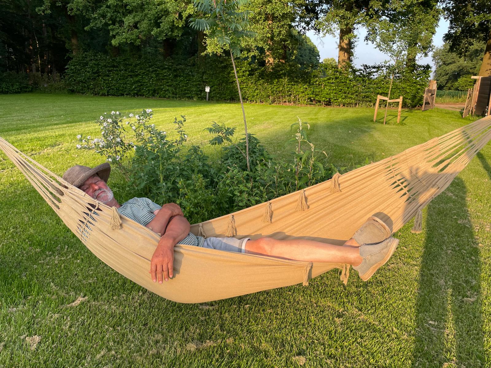 Older man with a hat relaxing in a beige hammock in a sunny backyard garden.