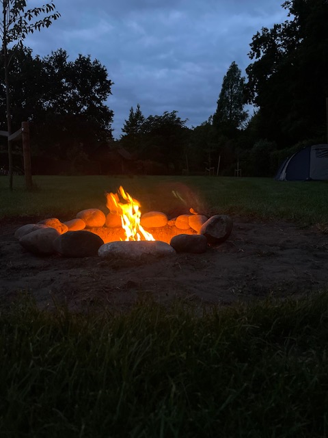 Campfire surrounded by stones in a campsite at dusk, with tents and trees in the background.